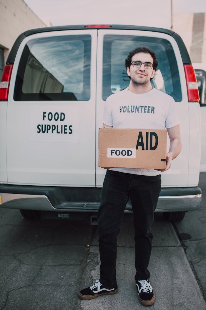 our-services-3 Volunteer standing in front of food supplies van with aid box, supporting community.